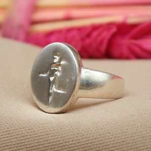 以下が含まれることがあります： テクスチャ加工が施された楕円形のトップを持つシルバーリング。リングはベージュの布地に置かれています。