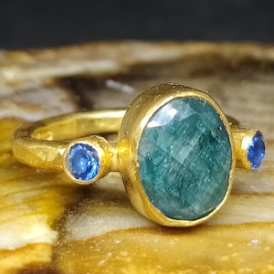 May include: A gold ring with a large oval green gemstone and two smaller blue gemstones. The ring is sitting on a piece of petrified wood.