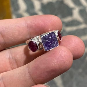 May include: A silver ring featuring a large, square, purple gemstone flanked by two smaller, round, red gemstones. The ring is held in a hand, showcasing the contrast of the polished metal and the vibrant colors of the stones. A unique piece of jewelry.