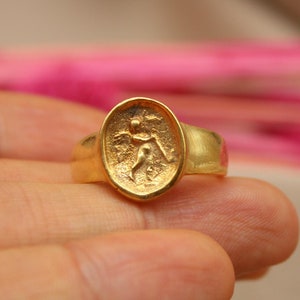 May include: A gold ring with a raised oval design featuring a detailed figure. The ring is worn on a finger.