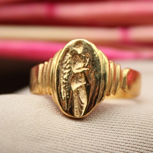Op de afbeelding: Een gouden ring met een gedetailleerd ontwerp van een figuur met vleugels. De ring is geplaatst tegen een roze achtergrond.