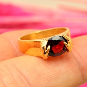 May include: A gold ring with a dark red gemstone. The ring is on a finger.