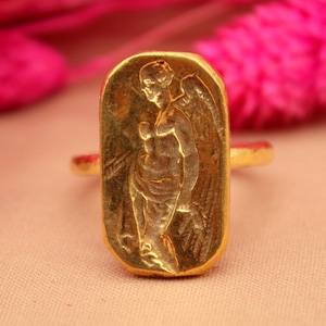 May include: A gold ring with a rectangular design featuring an embossed image of an angel with wings.