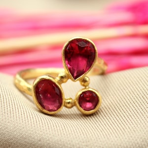 May include: A gold ring with three pink gemstones. The ring is set on a beige fabric background.