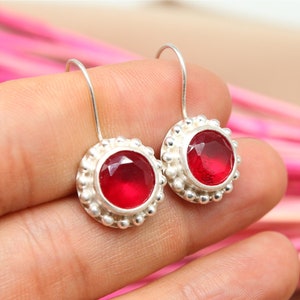 Op de afbeelding: Een paar zilveren oorhangers met rode edelsteen accenten. De oorhangers hebben een ronde, bezel-gezet edelsteen omgeven door een parelrand.