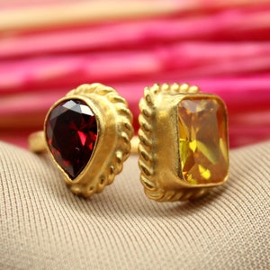 Op de afbeelding: Een gouden ring met twee edelstenen. De ring heeft aan de ene kant een druppelvormige rode edelsteen en aan de andere kant een vierkante gele edelsteen.