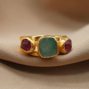 May include: Gold ring featuring three stones. The central stone is a large, square, green stone. The two smaller stones on either side are red.