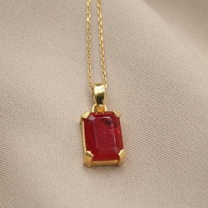Peut inclure: Un collier doré avec un pendentif rectangulaire rouge rubis. Le pendentif est serti dans un cadre doré et suspendu à une fine chaîne dorée. Le bijou est présenté sur un tissu de couleur neutre.
