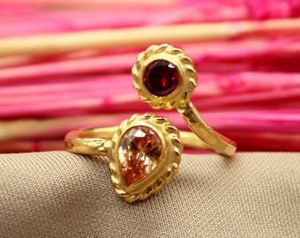 Puede incluir: Un anillo de oro con dos piedras preciosas. La piedra más grande es de forma de pera, de color naranja claro. La piedra más pequeña es redonda, de color rojo oscuro.