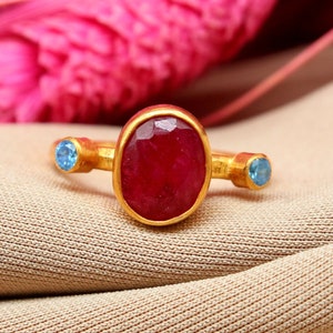 K&ouml;nnte beinhalten: Ein goldener Ring mit einem gro&szlig;en ovalen roten Edelstein und zwei kleineren blauen Edelsteinen an beiden Seiten.