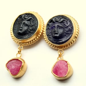 Könnte beinhalten: Goldene Ohrringe mit einem schwarzen und violetten Kamee eines Frauenkopfes und einem rosafarbenen Edelstein-Anhänger.