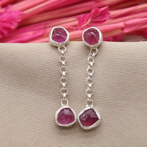 Op de afbeelding: Dangel oorbellen met ruwe, roze edelstenen gezet in zilver. De oorbellen hangen aan een delicate ketting, met een edelsteen aan de boven- en onderkant. De achtergrond is een neutrale beige kleur, met roze bloemen op de achtergrond.