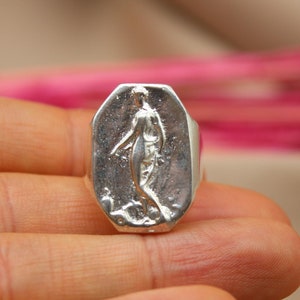 May include: A silver ring with a rectangular face featuring a sculpted figure. The ring is held in a hand, with a blurred background of pink and beige. The ring's design is a classical, artistic style.