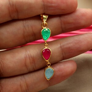 Può includere: Una collana con un ciondolo in oro con tre pietre preziose a forma di goccia. La pietra superiore è verde, la pietra centrale è rossa e la pietra inferiore è blu.