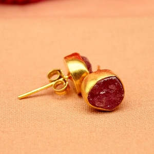 Op de afbeelding: Een paar goudkleurige oorstekers met een ruwe, donkerroze edelsteen. De oorbellen worden gepresenteerd op een perzikkleurig oppervlak, met een wazige fuchsia stof op de achtergrond. Ze hebben een pin en vlindersluiting.