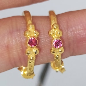 May include: A pair of gold hoop earrings with small pink gemstones. The earrings have a textured surface and are designed with a cluster of small beads.