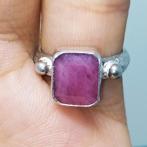 May include: A silver ring with a square, pink gemstone set in the center. The ring has two small silver balls on either side of the stone.