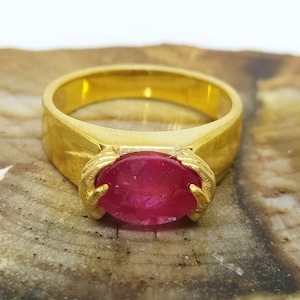 May include: A gold ring with a large, oval-shaped ruby gemstone set in the center. The ring is on a brown, textured surface.