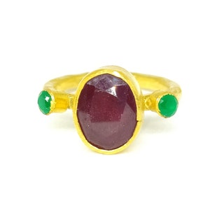 May include: A gold ring featuring a large oval ruby gemstone, flanked by two smaller green gemstones.