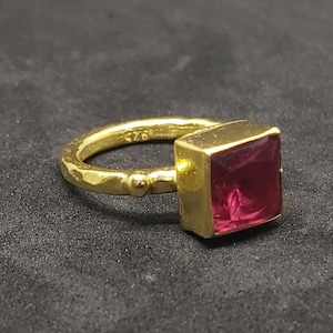 Könnte beinhalten: Ein goldener Ring mit einem quadratischen, rosafarbenen Edelstein. Die Ringband ist strukturiert und hat ein kleines Perlendetail.