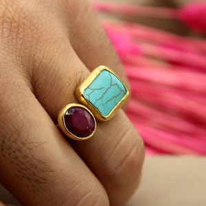 May include: A gold ring with a turquoise stone and a ruby stone. The ring is on a person's finger.