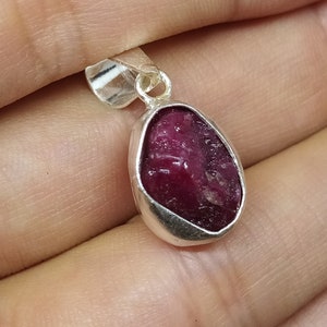 May include: A silver pendant featuring a raw, red ruby gemstone. The ruby is set in a silver bezel and has a natural, irregular shape.