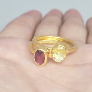 May include: Two gold rings with oval-shaped gemstones. The ring on the left has a red gemstone, and the ring on the right has a yellow gemstone.