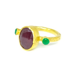 May include: A gold ring featuring a large, oval-shaped, dark red gemstone and two smaller, round, green gemstones.