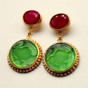 以下が含まれることがあります： ライオンのデザインが施された緑色のガラスのカメオと、トップに赤い宝石が付いた金色のイヤリングです。