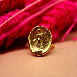 May include: A gold ring with an oval-shaped design featuring a raised image of a person. The ring is on a pink and beige background.