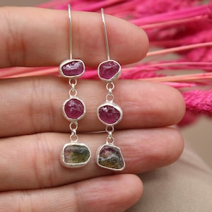 May include: A pair of silver dangle earrings with three pink and green gemstones. The earrings are hanging from a person's hand.