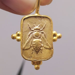 May include: Gold pendant charm featuring a detailed bee design. The charm is rectangular with rounded edges and has small gold beads on the sides.