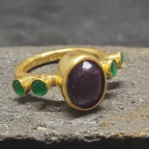 May include: A gold ring with a large purple gemstone and smaller green gemstones. The ring has a textured band.