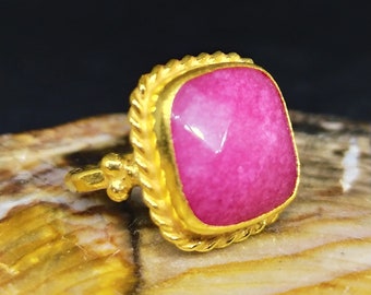 Può includere: Un anello d'oro con una pietra preziosa rosa incastonata in una lunetta quadrata. L'anello ha un design a corda attorno alla lunetta.