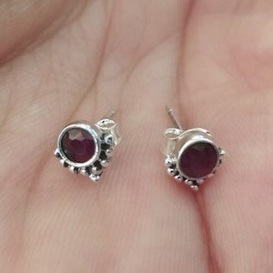 Op de afbeelding: Een paar zilveren oorbelletjes met een klein, rond, donkerrood edelsteentje in het midden van elke oorbel. De oorbelletjes hebben een delicaat, versierd ontwerp rondom het edelsteentje.