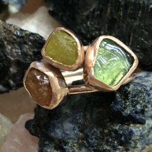 May include: A ring with three different colored gemstones set in copper. The gemstones are a yellow-green, a light green, and a brown.
