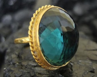 May include: A gold ring with a large, oval-shaped, faceted, teal blue gemstone. The ring has a rope-like design around the edge of the setting.