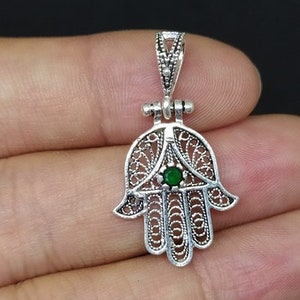 May include: Silver filigree hamsa hand charm with a green gemstone in the center. The charm has a loop at the top for attaching to a necklace or bracelet.