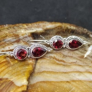 Op de afbeelding: Een paar zilveren oorbellen met rode edelsteen druppels. De oorbellen hebben een delicaat ontwerp met kleine kralen en een traanvormige edelsteen.