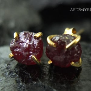 Op de afbeelding: Een paar gouden oorbel studs met rode robijn edelstenen. De robijnen zijn ruw en hebben een ruwe, natuurlijke textuur.