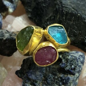 May include: Three gold rings with different colored gemstones. The rings feature a green, blue, and pink stone.