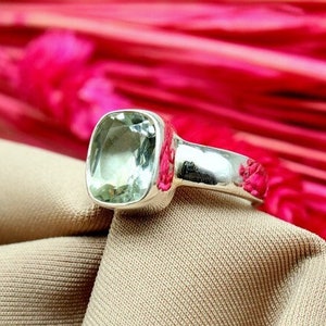 May include: A silver ring featuring a large, square-cut, pale green gemstone. The ring rests on a beige fabric, with a backdrop of bright pink, feathery flowers. The ring's band is polished and reflective, accentuating the gemstone's subtle colour.