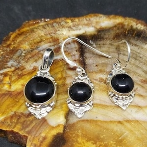 K&ouml;nnte beinhalten: Ein silbernes Anh&auml;nger- und Ohrring-Set mit schwarzen Onyx-Steinen. Die Steine sind in einem silbernen Filigran-Design gefasst. Der Anh&auml;nger und die Ohrringe befinden sich auf einem Hintergrund aus Naturstein.