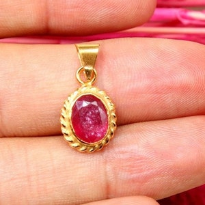 May include: A gold pendant with a red oval gemstone set in a gold bezel. The pendant has a rope-like design around the edge of the bezel.