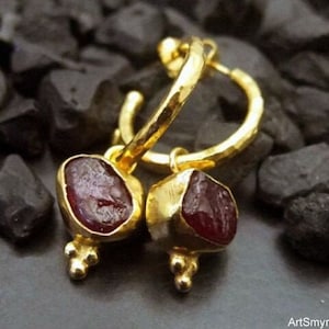 May include: Gold hoop earrings with dangling red gemstone drops. The hoops are hammered and have a textured finish. The gemstones are irregular in shape and have a deep red color.