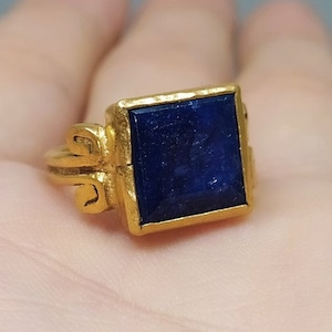 May include: A gold ring with a square blue gemstone set in a gold bezel. The ring has a decorative design on the band.