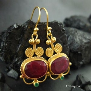 May include: Gold earrings with a red gemstone and green accents. The earrings have a delicate, intricate design.