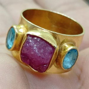 May include: A gold ring with a large, faceted, pink ruby stone and two smaller, oval, blue topaz stones.