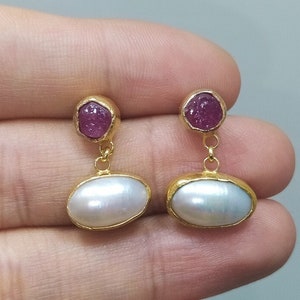 Op de afbeelding: Gouden oorbellen met een roze edelsteen en een witte parel hanger.