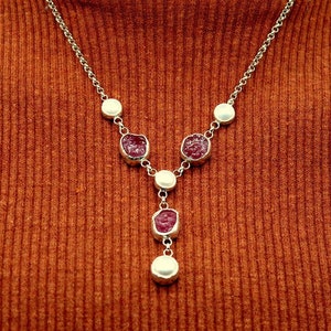 May include: A silver necklace with a Y-shaped design features five round, white pearl-like beads and four red, faceted gemstone pendants.